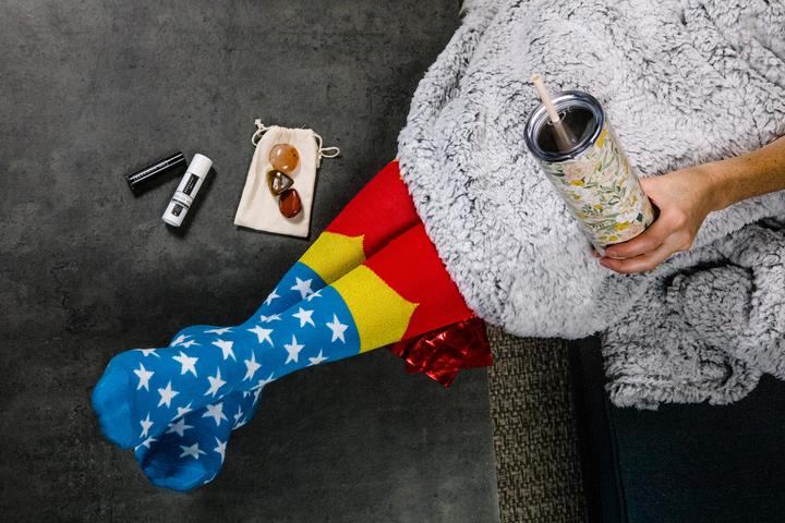 Image of a woman using the contents of the cancer warrior gift basket. Wearing wonder woman socks, drinking from waterbottle, while covered by an amazing fuzzy blanket healing stones and other gift items are on the floor beside her.