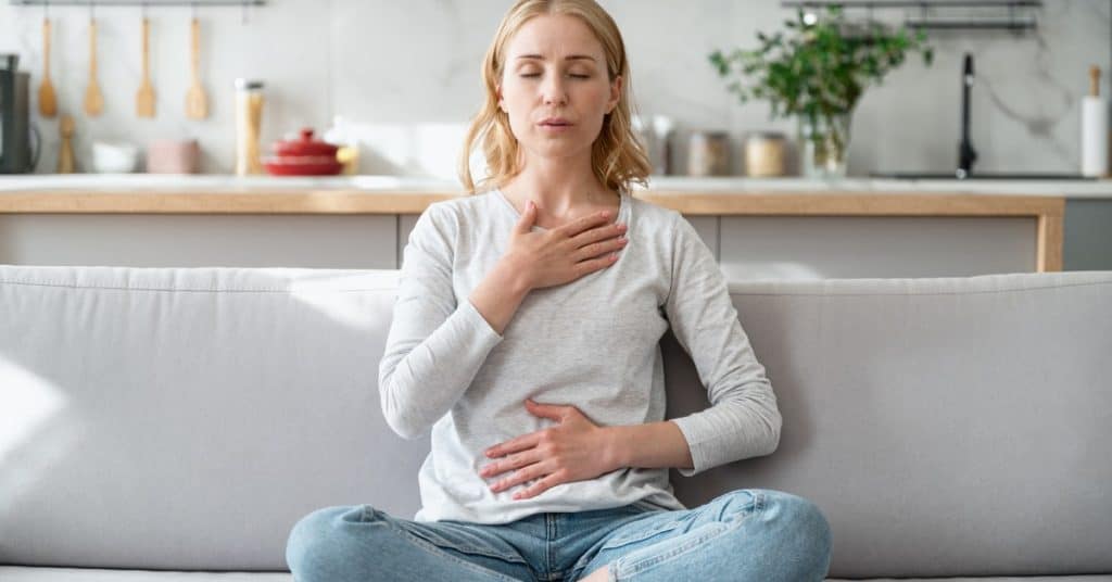Woman on her courch practicing deep breathing. One hand on her belly, one hand on her chest.